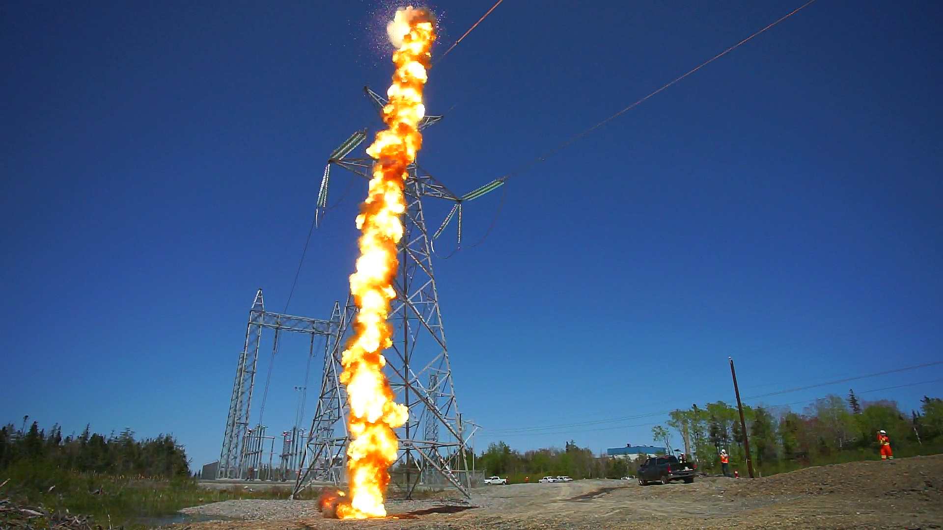 A line of flame erupting from a power transmission tower against a clear blue sky.